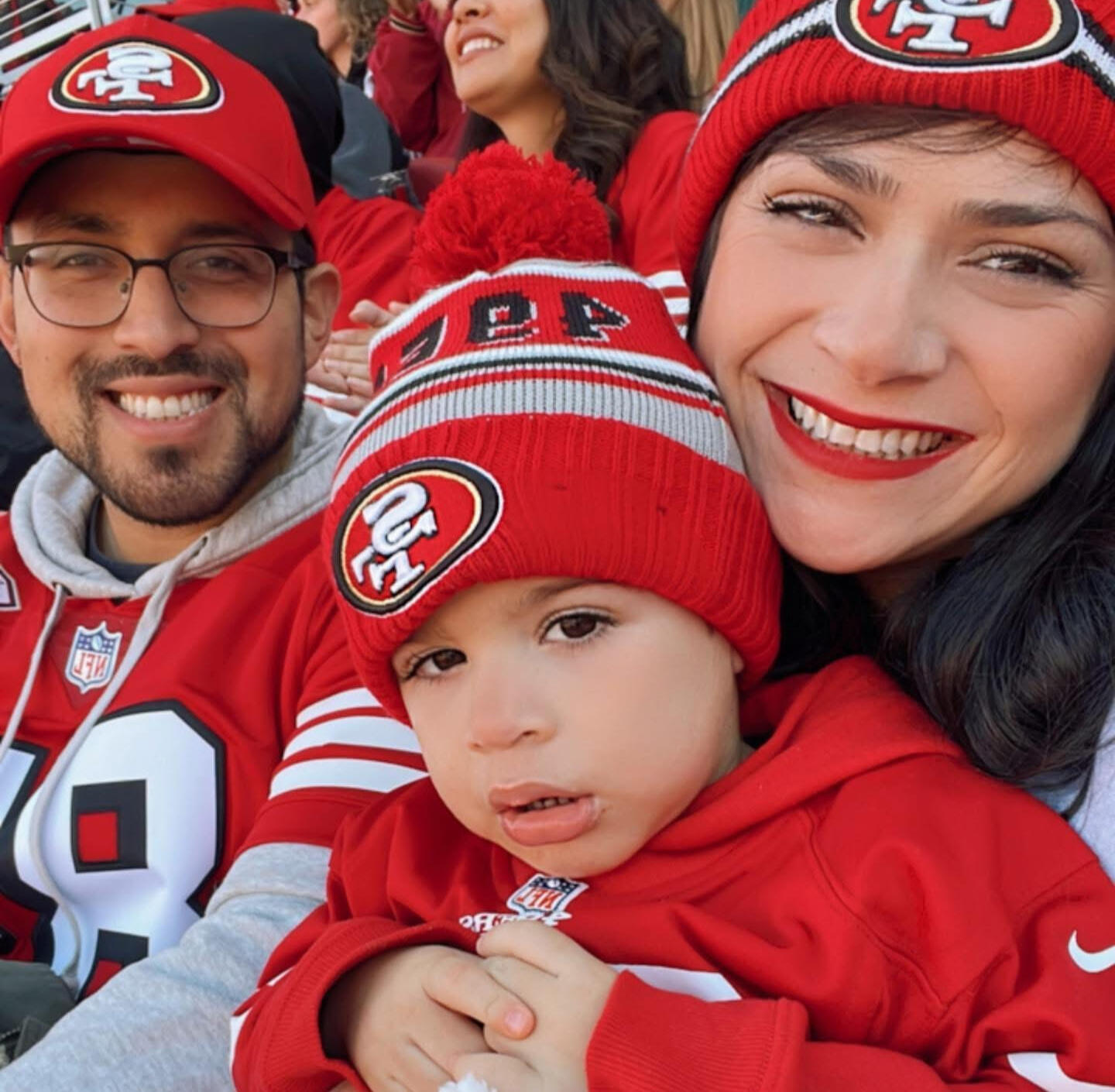 Football Game A selfie of the founder, Michael Cuellar, with his wife and son sitting in the stands of a football game wearing San Francisco 49er jerseys and hats.